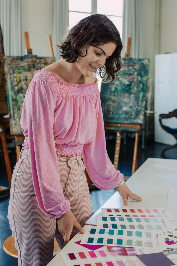 A female student with dark hair in a pink blouse stands in front of a workbench on which various cards with colour schemes are spread out in front of her.