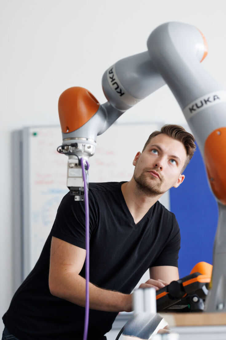 Student in a dark t-shirt operates the arm of a robot by remote control.