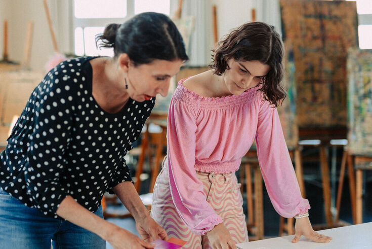 A student in a pink blouse bends over design drafts with her lecturer. In the background you can see colourful canvases on easels.