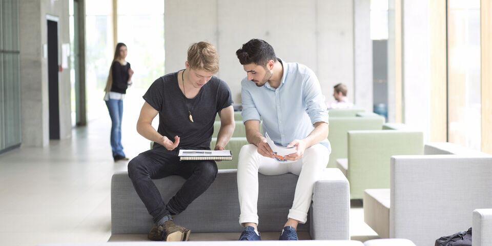 A blonde student in a black T-shirt and a dark-haired student in a light blue shirt bend over a notepad and talk.