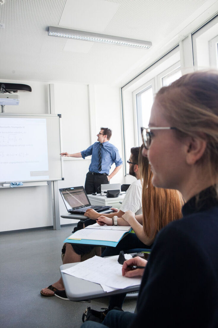 Students are sitting in the seminar room; a lecturer stands in front of the whiteboard and points to it. 